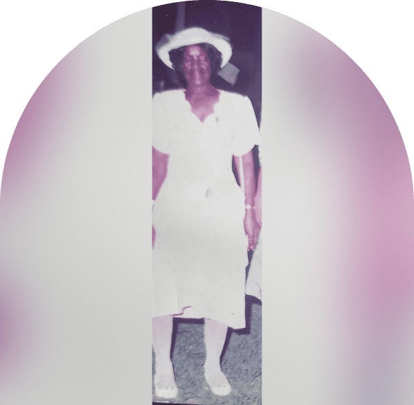 An elderly woman in a white dress and hat stands smiling.